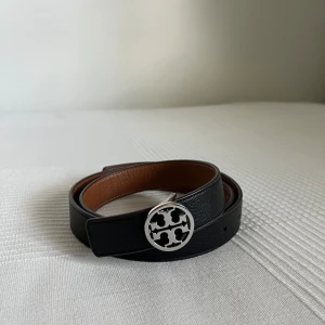 Svart skärp från Tory Burch i skinn - Snyggt svart bälte från Tory Burch i äkta skinn med brunt foder och en rund, silverfärgad logga som spänne. Perfekt accessoar för att lyfta din outfit med en stilren touch och exklusiv känsla. Bältet är i storlek M. Säljer då den inte komme till användning. Inköpt på Nordiska Kompaniet i Göteborg. 