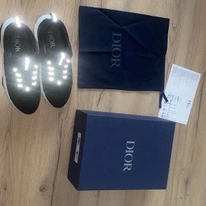 Dior B22 - Svarta Dior sneakers med vit sula och reflekterande detaljer på snörningen. Ovandelen är i mesh och skinn för en modern look. Snygg logga på sidan och insidan. Perfekta för dig som gillar exklusiva och trendiga sneakers.