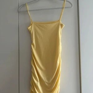 Gul kort klänning med drapering - Snygg gul kort klänning med smala axelband och draperad effekt längs sidorna. Klänningen har en enkel, figurnära passform och är perfekt för varma dagar. Materialet känns mjukt och stretchigt