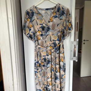 Blommig långklänning från Second Female - Klänning med blommigt mönster i beige, blått och gult från Soyaconcept. . Klänningen har v-ringning, korta ärmar och knytskärp i midjan. Material viscose. . Snygg och flowy siluett som ger en chill vibe.