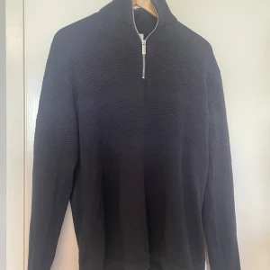 Mörkblå stickad half zip-tröja Selected - Svart stickad tröja från Selected Homme i storlek M. Tröjan har en snygg half zip-dragkedja och strukturstickade partier över bröst och ärmar. Hög krage och långärmad modell, perfekt för lager-på-lager under kyliga dagar.