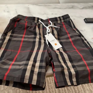 Rutig shorts med snörning - Snygga rutiga shorts med svart, beige och röda ränder. De har elastisk midja och vit snörning framtill för skön passform. Perfekta för sommaren och har en klassisk streetstyle-vibe. Lätta och bekväma, passar till många olika outfits.