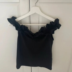 Svart offshoulder topp med volang - Supersnygg svart offshoulder topp med bred volang runt hela ringningen. Toppen är ärmlös och har en tight passform som framhäver axlarna. Perfekt för dig som vill ha en trendig och stilren look med lite extra edge.