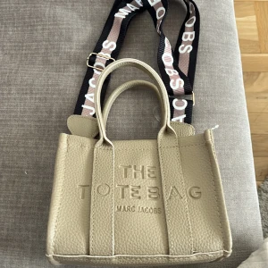Beige The Tote Bag från Marc Jacobs - Snygg beige handväska från Marc Jacobs i präglat skinn med texten THE TOTE BAG framtill. Väskan har både handtag och en bred axelrem med logga, samt dragkedja upptill. Perfekt storlek för vardagens essentials och ger en cool vibe till din outfit. Går att ta bort banden så man kan variera hur man vill ha den! 