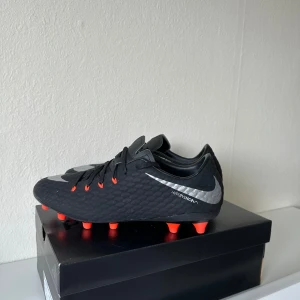 Nike Hypervenom svart fotbollsskor - Svarta Nike Hypervenom fotbollsskor med silverfärgad swoosh och röda dobbar. Ovandelen har en strukturerad yta och snörning framtill. Skorna är tillverkade i syntetmaterial och har en klassisk låg profil som passar perfekt för spel på gräs.