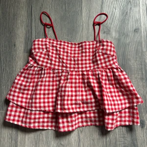 Rutig röd och vit volangtopp - Supersöt rutig topp i rött och vitt med tunna axelband och dubbla volanger. Perfekt för sommaren med sin luftiga känsla och trendiga look. Toppen har en croppad passform och smala axelband som jag knutit men kan öppnas. Ger en lekfull och retro vibe.