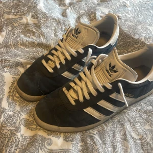 Adidas Gazelle sneakers i mörkblå mocka - OBS! De blåa snören medföljer också:) Säljer ett par klassiska Adidas Gazelle sneakers i mörkblå mocka med vita detaljer och snörning. Skorna har de ikoniska tre ränderna på sidan, vit sula och Adidas-logga på häl och plös. Även öppen för byte! 