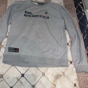 Grå Sail Racing sweatshirt - Snygg grå sweatshirt från Sail Racing med trycket 'Sail Antarctica' på bröstet och en liten patch nertill. Tröjan har rund hals, långa ärmar och är gjord i mjuk bomull. Perfekt för dig som gillar stilren och sportig look.