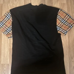 Burberry svart t-shirt - Svart t-shirt från Burberry med klassiska rutiga korta ärmar i beige, rött, vitt och svart. T-shirten har en rak passform och rund halsringning. Perfekt för dig som vill ha en stilren look med en touch av lyxig streetstyle.