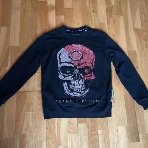 Svart Philipp Plein tröja med döskalle M - Svart långärmad tröja från Philipp Plein med en stor döskalle framtill i silver och rött med glittriga paljettdetaljer. Rund halsringning och ribbade muddar. Märkeslogga på bröstet och etikett i sidan. Perfekt statement piece för dig som gillar edgy stil. FINNS INTE ATT KÖPA NÅGONSTANS !!!