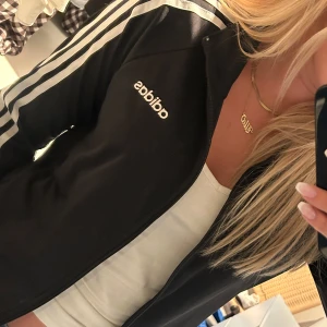 Svart Adidas vindjacka med vita ränder - Svart vindjacka från Adidas med klassiska vita ränder längs ärmarna och vit Adidas-logga på bröstet. Jackan har dragkedja framtill och är tillverkad i ett lätt och sportigt material. Perfekt för en sportig och avslappnad stil.