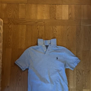 Ljusblå pikétröja från Ralph Lauren - Säljer en ljusblå pikétröja från Polo Ralph Lauren i bomull. Klassisk modell med korta ärmar, krage och knappar framtill. Broderad logga på bröstet och slim fit-passform. Perfekt för en clean och stilren look.