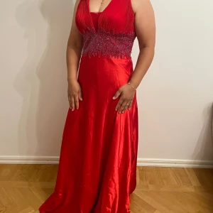 Röd långklänning med pärldetaljer - Snygg röd långklänning med djup v-ringning och glittrande pärldetaljer i midjan. Överdelen är plisserad och kjolen har ett elegant fall. Perfekt för bal eller fest. Materialet har en glansig finish som ger en lyxig känsla. Använd endast en kväll. 