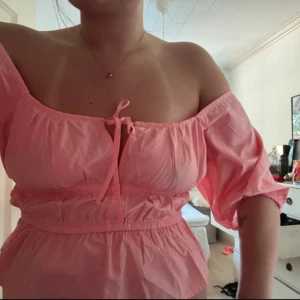 Rosa offshoulder blus med knyt - Supersöt rosa blus med offshoulder och puffiga ärmar. Toppen har smock i midjan och ett knyt framtill som ger en romantisk vibe. Perfekt för dig som gillar en feminin och trendig look.