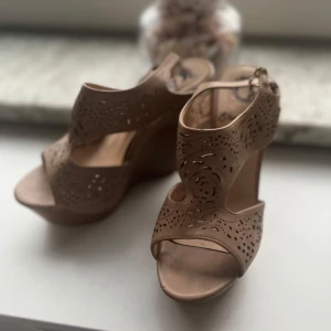 Beige kilklackade sandaler med stjärnberlock - Snygga beige sandaler med hög kilklack och öppen tå. Skorna har utskurna detaljer i lädret och en söt stjärnberlock på remmen. Perfekta för sommarens alla tillfällen när du vill sticka ut lite extra.