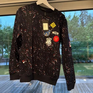 Svart Dsquared2 sweatshirt med badges - Cool svart sweatshirt från Dsquared2 med färgstänk i rött och vitt samt flera broderade badges på bröstet. Tröjan har rund hals, ribbade muddar och är långärmad. Perfekt statement-piece för dig som gillar streetstyle och unika detaljer.