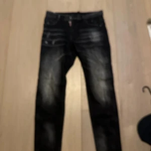 Svarta jeans från Dsquared2, strl 33 - Säljer ett par svarta jeans från Dsquared2 med coola slitningar och tydliga detaljer. Jeansen har klassisk femficksdesign, Dsquared2-logga bak och snygg tvättad look. Perfekta för dig som vill sticka ut med ett par trendiga jeans.