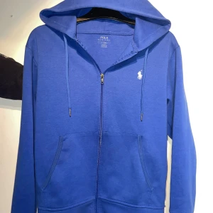 Blå hoodie från Polo Ralph Lauren - Snygg blå hoodie från Polo Ralph Lauren med dragkedja och klassisk vit logga på bröstet. Tröjan har huva med snörning och två stora fickor framtill. Perfekt för dig som gillar en stilren och sportig look.