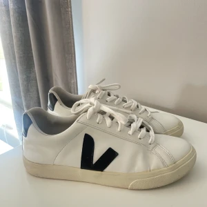 Vita Veja sneakers med svarta detaljer - Snygga vita sneakers från Veja med svart V-logga på sidan och svart hälparti med vit Veja-text. Klassisk låg modell med snörning och platt sula. Tillverkade i skinn och passar perfekt till en clean och stilren look.