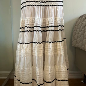 Vit långklänning från Maje med spets - Lång maxikjol från Maje i vitt bomullstyg med broderade spetsdetaljer och svarta vågiga ränder. Klänningen har en boho-stil och dragkedja vid sidan.