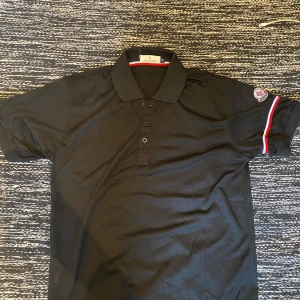 Svart Moncler pikétröja - Svart pikétröja från Moncler med klassisk krage och knappar framtill. Ärmarna har snygga röd, vit och blå ränder samt broderad Moncler-logga på vänster ärm. Storlek S