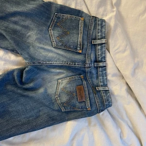Blå bootcut jeans från Wrangler - Snygga blå jeans från Wrangler med klassiska kontrastsömmar och bootcut passform. Jeansen har fem fickor, bälteshällor och en läderpatch med logga på bakfickan. Perfekta för dig som gillar en retro vibe och vill ha ett par jeans med  utsvängda ben.