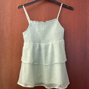 Ljusgrön volangtop från Name It - Linne strl 146-152. Supersöt ljusgrön linnetopp från Name It med smala axelband och smock upptill. Toppen har två lager volanger i ett tunt, prickigt tyg som ger en luftig och somrig känsla. Perfekt för dig som gillar en romantisk och lätt stil.