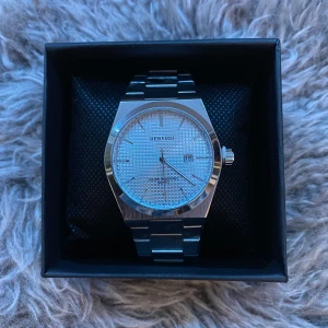 Silverfärgad klocka från Denvosi - Stilren klocka från Denvosi i silverfärgat metall med länkarmband. Urtavlan är ljus med rutmönster och har datumvisning vid klockan tre. Klassisk design med tydliga index och kvartsverk. Perfekt accessoar för dig som gillar en clean look.