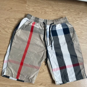 Burberry shorts - Snygga shorts med klassiskt rutigt mönster i beige, svart, vitt och röda detaljer. De har elastisk midja med snörning och sidofickor, samt en bakficka. Perfekta för sommaren och riktigt sköna att bära. Materialet känns lätt och luftigt.