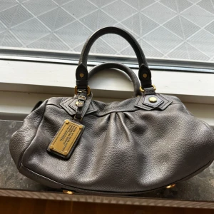 Grå skinnväska Marc by Marc Jacobs - Snygg grå handväska från Marc by Marc Jacobs i äkta skinn med guldfärgade metalldetaljer och klassisk logga på bricka framtill. Denna Marc Jacobs-väska är i gott skick och i storlek small. Den har en avtagbar axelrem, vilket gör att den kan bäras både som handväska och axelväska för en mångsidig stil. 100 % koskinn.