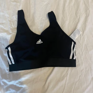 Svart sporttopp från Adidas M - Svart, vadderad sport-bh från Adidas med vita ränder på sidorna och Adidas-logga framtill. Tillverkad i ett stretchigt material, perfekt för träning. Bred resår nedtill och smala axelband för extra stöd. Använd fåtal gånger