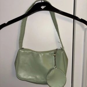 Ljusgrön axelväska med rund miniväska - Snygg ljusgrön axelväska i syntetmaterial med en rund miniväska som hänger i en kedja. Väskan har dragkedja upptill och bred axelrem. Väskan har en avdragsbar plånbok.