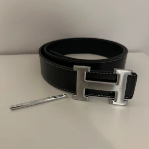 H bälte - Svart bälte med ikoniskt H-spänne i silverfärgad metall. Bältet är gjort i skinn och har vita kontrastsömmar längs kanterna. Perfekt accessoar för att lyfta din outfit med en lyxig touch.
