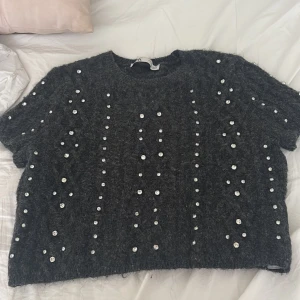 Grå stickad tröja med pärlor från Zara - Mörkgrå stickad tröja från Zara med korta ärmar och rund hals. Tröjan är dekorerad med glittrande pärlor i rader över hela plagget, vilket ger en unik och trendig look. Perfekt för dig som vill sticka ut lite extra.