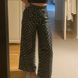Svarta prickiga vida kostymbyxor - Svarta byxor med vita prickar, hög midja och visa ben. Byxorna har dragkedja i sidan och knappstängning. Materialet känns lätt och svalt. Handsydda. Aldrig använda.