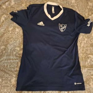 Blå IFK fotbollströja från Adidas - Snygg marinblå fotbollströja från Adidas med IFK Uppsala-logga på bröstet och stor IFK-tryck på ryggen. Tröjan har vit V-ringad krage och korta ärmar. Tillverkad i lätt och funktionellt material som passar perfekt till träning eller match.