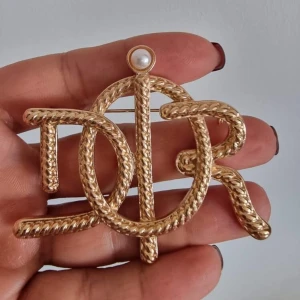 Guldfärgad Dior brosch med pärla - Snygg brosch från Dior i guldfärgad metall med flätad struktur och en vit pärldetalj på toppen. Broschen har de ikoniska Dior-bokstäverna i en cool, chunky design som verkligen sticker ut.