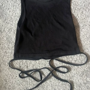 Svart ribbad linnetopp med snörning - Snygg svart ribbad linnetopp med smala axelband och långa snören i nederkant som kan knytas runt midjan. Perfekt för att skapa en trendig look och passar till både jeans och kjol. Toppens material är mjukt och stretchigt för en bekväm passform.