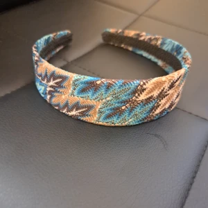 Mönstrat diadem i blått och beige - Snyggt diadem med bred form och coolt mönster i blått, beige och brunt. Tygklädd yta med grafiskt, nästan etnoinspirerat mönster som ger en unik touch till din look. Perfekt accessoar för att lyfta vilken outfit som helst.