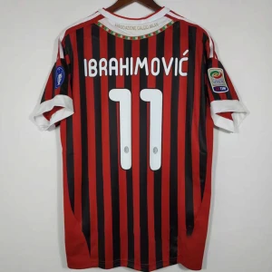 AC Milan Ibrahimović matchtröja - Snygg AC Milan fotbollströja med Ibrahimović och nummer 11 på ryggen. Tröjan är röd och svart med vertikala ränder, vita detaljer vid ärmslut och krage samt officiella Serie A- och klubbmärken på ärmarna. Perfekt för fans av Zlatan och italiensk fotboll.