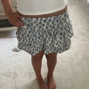 Vit och blå mönstrad kjol från Zara - Supersöt kortkjol från Zara i vitt med blått mönster. Kjolen har en luftig passform, resår i midjan och volangig nederkant. Perfekt för varma dagar och ger en chill och fräsch vibe till din outfit.