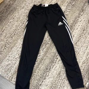 Svarta Adidas träningsbyxor - Svarta träningsbyxor från Adidas med vita ränder längs sidorna och logga på låret. Byxorna har elastisk midja med snörning och dragkedjefickor 