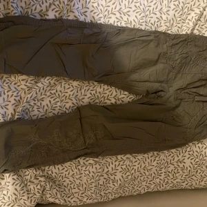 Olivgröna cargopants med broderi - Säljer ett par olivgröna cargopants med snygga broderade detaljer på ena benet. Byxorna har flera fickor och avslappnad passform, perfekta för en chill och trendig look. Materialet känns lätt och skönt mot huden.