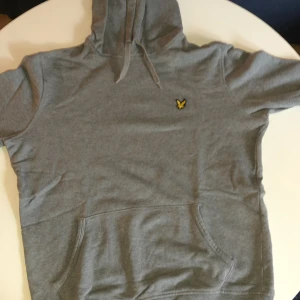 Grå hoodie från Lyle & Scott - Snygg grå hoodie från Lyle & Scott med klassisk gul logga på bröstet. Tröjan har huva med snörning och en stor magficka framtill. Perfekt för dig som gillar en enkel och stilren look.