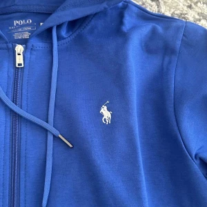 Blå hoodie från Polo Ralph Lauren - Snygg blå hoodie från Polo Ralph Lauren med dragkedja och huva. Klassisk vit broderad logga på bröstet och praktiska fickor framtill. Tillverkad i mjuk bomull, perfekt för chill dagar. Stilsäker och enkel att matcha med jeans eller joggers.