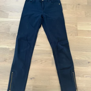 Svarta byxor Holy & Whyte med dragkedjor - Svarta byxor från Holy & Whyte by Lindex i stretchig bomull med skinny fit. Snygga detaljer med guldiga dragkedjor vid bensluten och resår i midjan för extra komfort. Klassisk femficksmodell, perfekta till sneakers eller boots.