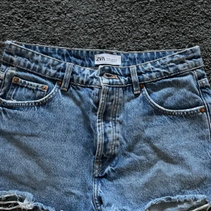 Blå jeansshorts från Zara - Snygga blå jeansshorts från Zara med fransig kant och klassisk femficksdesign. Shortsen har normal passform och är perfekta för sommaren. Tillverkade i slitstarkt denim med dragkedja och knapp framtill.