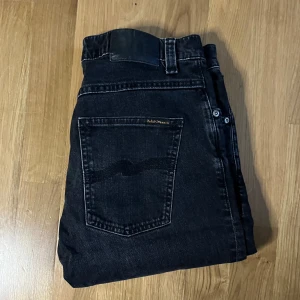 Nudie Grim Tim jeans - Säljer ett par svarta Nudie Jeans Grim Tim i modellen Concrete Black. Jeansen har klassisk femficksdesign, raka ben och subtila slitningar. Materialet är 99% bomull och 1% elastan för lite stretch. Perfekta för dig som gillar en clean och stilren look. Midja ca 40cm. Jag skickar ej bild när jag bär plaggen och jag vet ej vilken längd och vikt de passar då jag inte prövar kläderna jag säljer, så för storlek hänvisar jag till måtten och passform att man googlar på modellnamn etc 😃