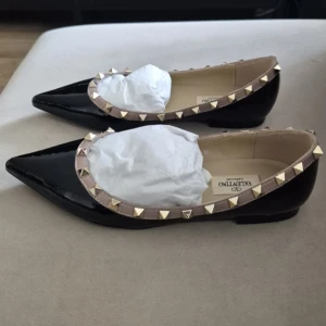 ballerinaskor svart - Snygga ballerinaskor i svart lackat skinn med spetsig tå. Skorna har beige kant med ikoniska guldiga nitar runtom för en edgy och lyxig look. Perfekta för dig som vill sticka ut med stilrena detaljer.