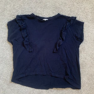 Marinblå topp med volangdetaljer - Säljer en marinblå topp från Marine i mjuk bomull. Toppen har korta ärmar och volangdetaljer som går från axlarna ner över framsidan. 